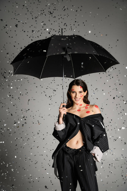 веселая женщина с красными отпечатками губ на теле в черной одежде и стоящая с зонтиком под падающими конфетти на сером фоне - Фото, изображение