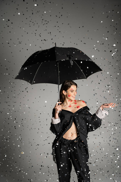 happy woman with red kiss prints on body wearing trendy clothes and standing under black umbrella and confetti on grey  - Foto, Bild