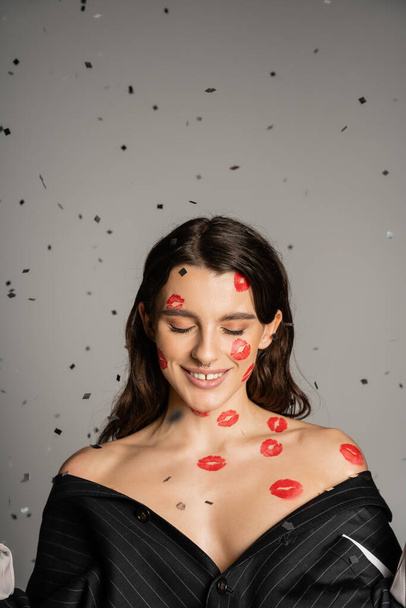 brunette woman with red kiss prints on face and bare shoulders smiling with closed eyes on grey background - Foto, immagini