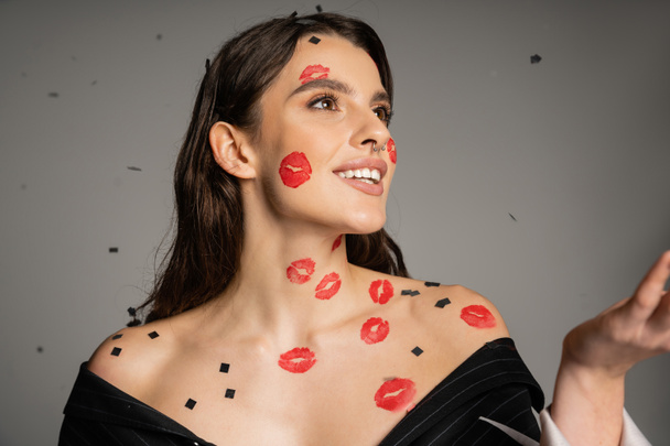 cheerful woman with red lip prints and confetti on face and bare shoulders looking away on grey background - Foto, Bild