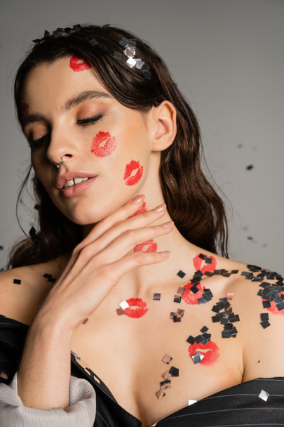 sensual woman with red kiss prints and shiny confetti on face and naked shoulders posing with closed eyes on grey  - Fotografie, Obrázek