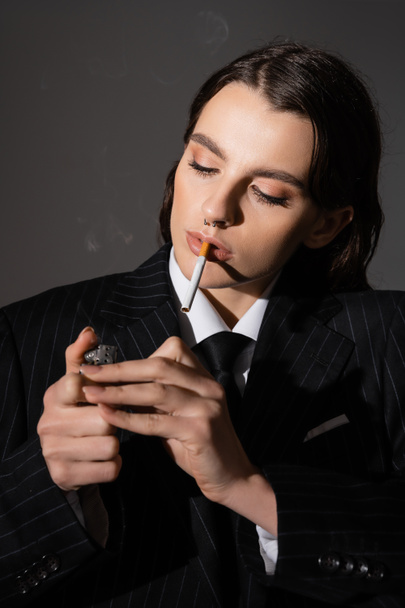 portrait of young model in formal wear lighting cigarette isolated on dark grey - Foto, afbeelding