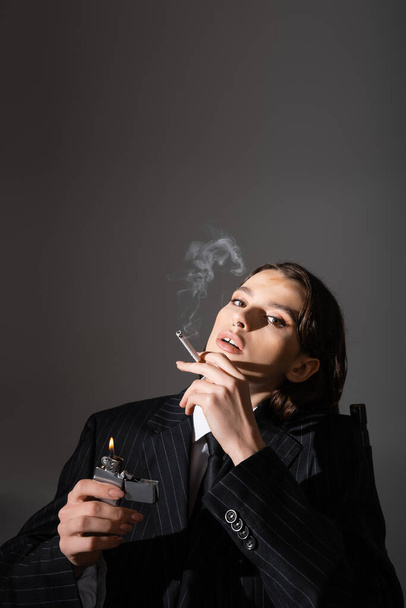 sensual woman in black striped suit holding lighter and smoking isolated on grey with copy space - Foto, Imagem