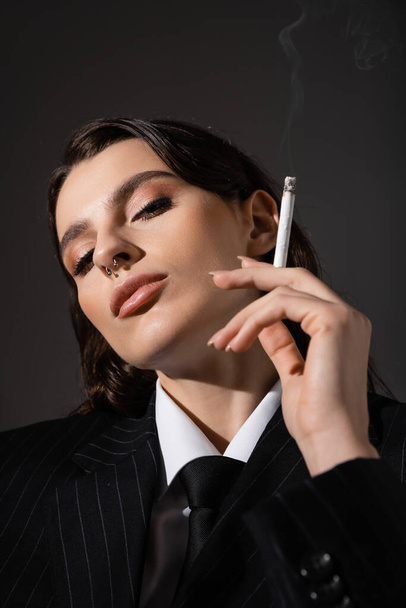 портрет молодої брюнетки в елегантному вбранні, що тримає сигарету і дивиться на камеру ізольовано на темно-сірому
 - Фото, зображення