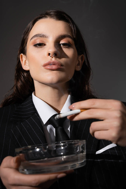 portrait of fashionable woman with makeup and piercing holding ashtray and cigarette isolated on dark grey - Fotografie, Obrázek