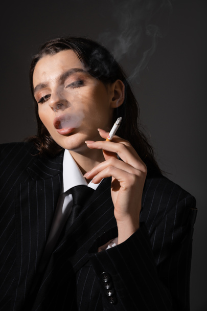 sensual brunette woman in black blazer and tie smoking isolated on dark grey - Zdjęcie, obraz