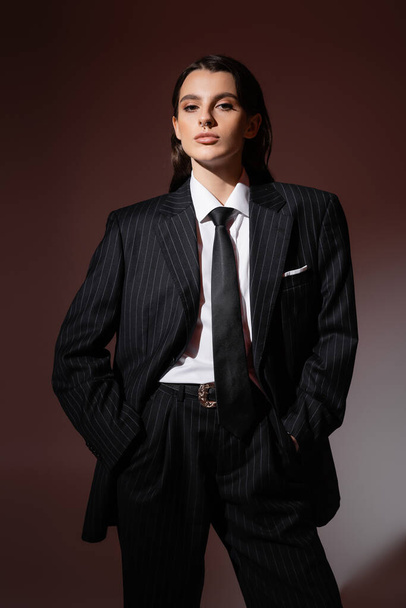 trendy woman in black striped pantsuit and tie standing with hands in pockets and looking at camera on brown background - Foto, Imagem