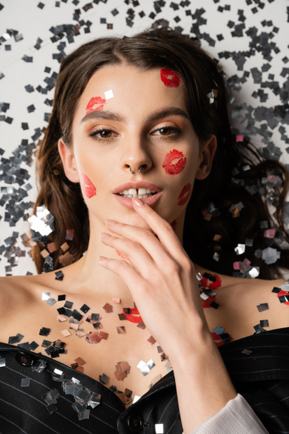 top view of sensual woman with makeup and red kiss prints touching lip near silver confetti on grey background - Foto, Imagen