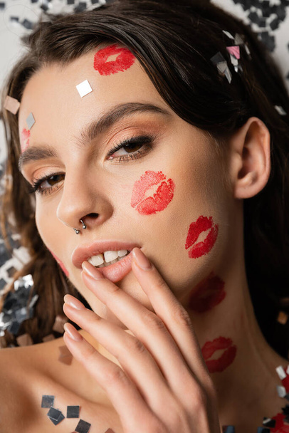 portrait of sensual woman with makeup and red kiss prints touching lips and looking at camera near silver confetti on grey  - Photo, Image