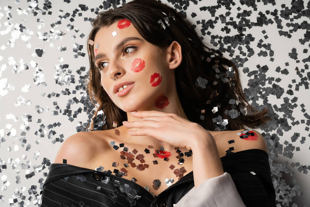 top view of young brunette woman with red lip prints looking away near sparkling silver confetti on grey background - Foto, imagen
