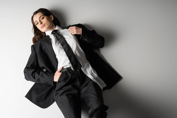 high angle view of brunette woman in white shirt and black striped suit lying with hand in pocket on grey background - Φωτογραφία, εικόνα