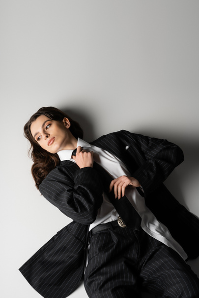 high angle view of brunette woman in black oversize suit touching tie and looking away while lying on grey background - Fotografie, Obrázek