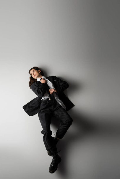 high angle view of model in elegant formal wear and rough boots touching black tie while lying on grey  - Foto, Bild