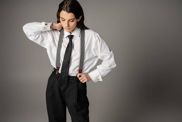 young and stylish woman in oversize shirt and black striped pants touching neck on grey background - Foto, afbeelding