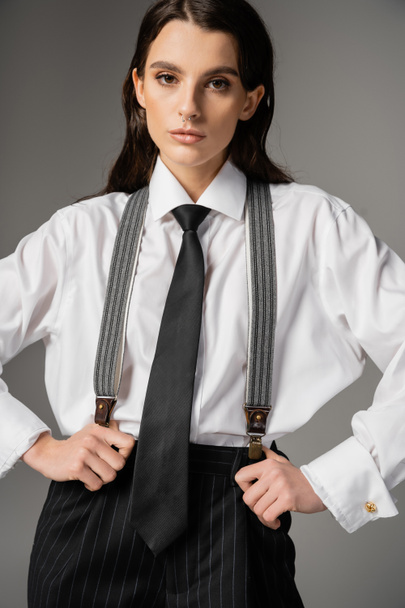 brunette woman in white oversize shirt and tie holding black pants with suspenders and looking at camera isolated on grey - Fotoğraf, Görsel