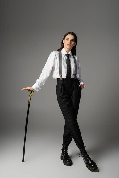 full length of young woman in elegant clothes and rough leather boots posing with walking cane on grey background - Foto, imagen