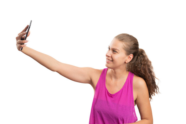 Female person making smiling friendly expression taking selfie photo using smartphone wearing fitness clothing pink tanktop isolated on white studio background - Photo, image