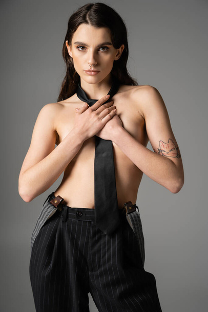 shirtless tattooed woman in black tie and black striped pants covering breast and looking at camera isolated on grey - Photo, image