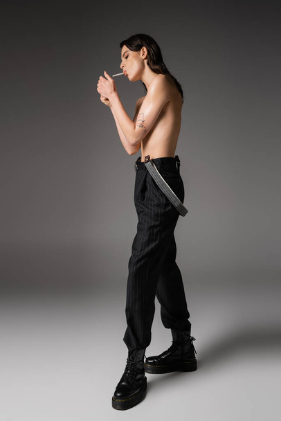 full length of shirtless woman in black trousers and rough boots lighting cigarette on grey background - Foto, afbeelding