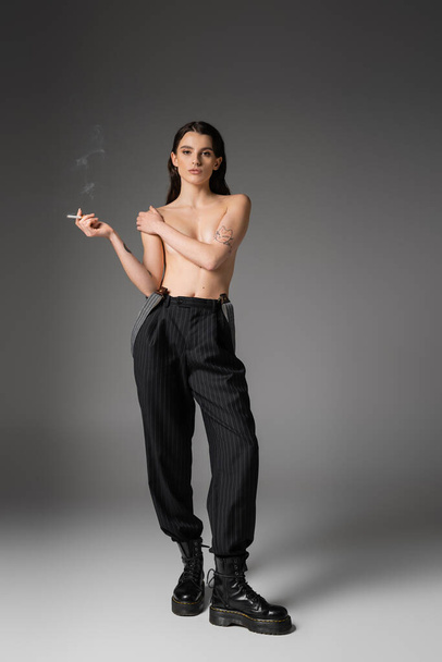 full length of shirtless and sexy woman in black pants and rough boots posing with cigarette and looking at camera on grey  - Foto, Bild