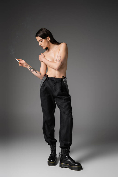 full length of shirtless tattooed woman in trendy pants and laced-up boots covering breast and holding cigarette on grey  - Photo, Image
