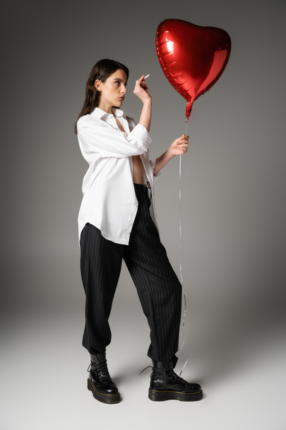 full length of woman in white shirt and black pants with rough boots holding cigarette near red heart-shaped balloon on grey  - Zdjęcie, obraz