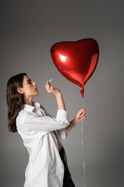 side view of brunette woman in white shirt holding cigarette near red heart-shaped balloon on grey  - Foto, afbeelding
