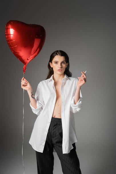 stylish woman with red heart-shaped balloon and cigarette posing in white unbuttoned shirt on grey background - Foto, Imagen