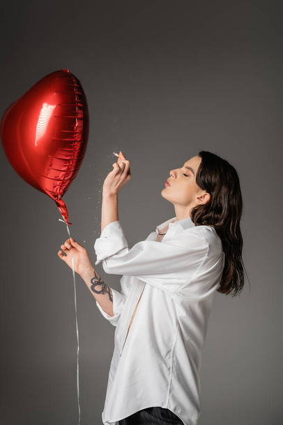 side view of young tattooed woman in white shirt holding cigarette near red festive balloon on grey  - Fotografie, Obrázek
