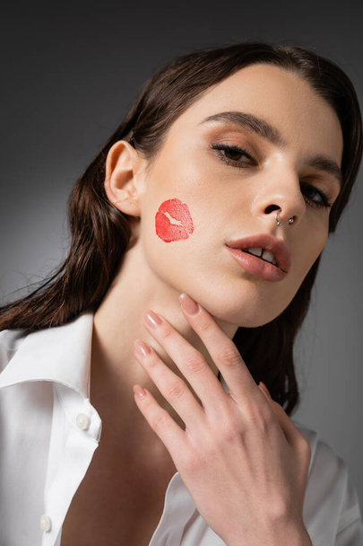 portrait of brunette woman with red kiss print on face touching neck and looking at camera on grey background - Foto, afbeelding