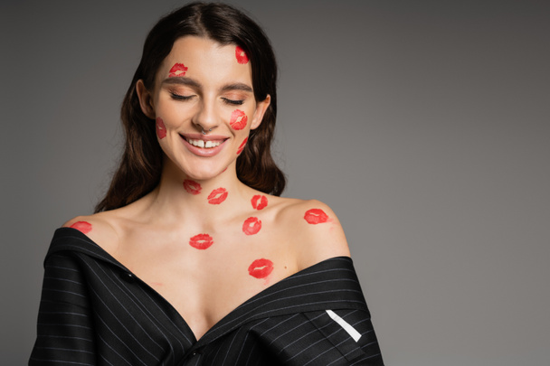 happy young woman with red lipstick marks on face and bare shoulders smiling with closed eyes isolated on grey - Foto, imagen