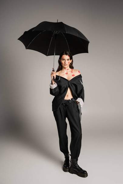 full length of sexy woman with red kiss prints wearing oversize suit while holding umbrella on grey background - Foto, Bild