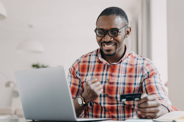 E-banking, e-commerce. African american man use e banking service at laptop. Black guy satisfied by easy secure payment, receiving benefits as loyal client, paying online, holding plastics credit card - Foto, imagen