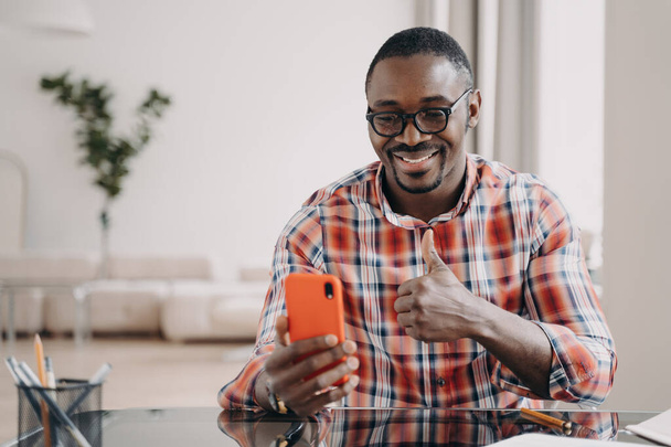 Happy african american man showing like thumb up hand, holding smartphone smiling. Pleased black guy looking at phone screen, showing positive feedback gesture, chatting by video call, sitting at desk - Foto, Bild