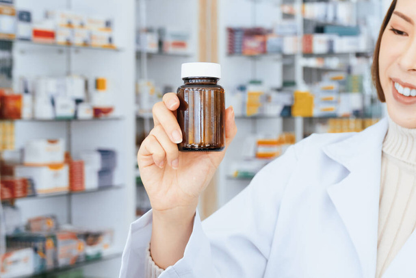 Closeup young affable pharmacist show qualified medicine pill bottle mockup product for copyspace at pharmacy. Druggist working with her diary job at drugstore, medicine box on shelf at background - Photo, Image