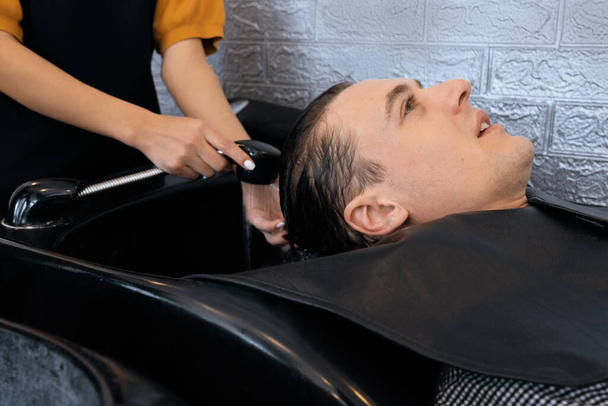 Detailní kavkazský muž, který si myje vlasy profesionálním kadeřníkem se šamponem, se cítí uvolněně a pohodlně. Kvalifikovaná kadeřnice se svým holičstvím, kadeřnictví poskytující koncept péče o vlasy. - Fotografie, Obrázek