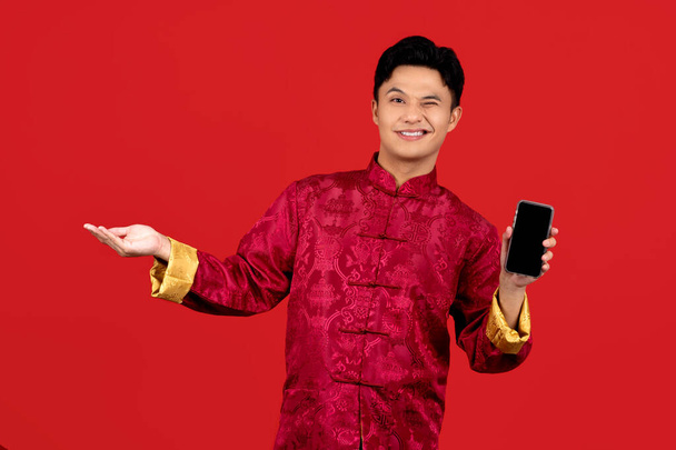 Счастливый молодой азиат в красном Чхонсам Кипао показывает мобильный телефон изолирован на красном фоне. - Фото, изображение