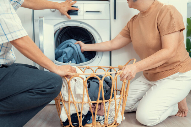 Senior couple working together to complete their household chores at the washing machine in a happy and contented manner. Husband and wife doing the usual tasks in the house. - Фото, изображение