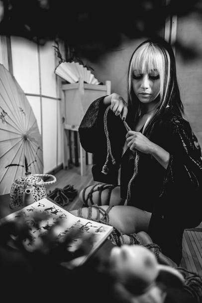Beautiful, slim, young and sexy girl with blonde locks, small and sexy black kimono and panties, and shibari ties and knots on the body, in a traditional japanese room (in black and white) - Photo, image