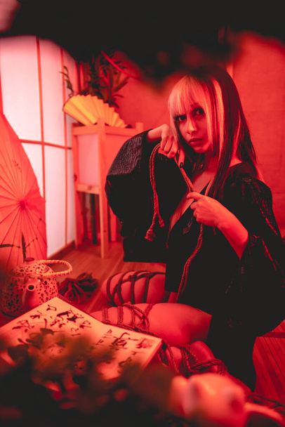 Beautiful, slim, young and sexy girl with blonde locks, small and sexy black kimono and panties, and shibari ties and knots on the body, in a traditional japanese room with red light - Photo, Image