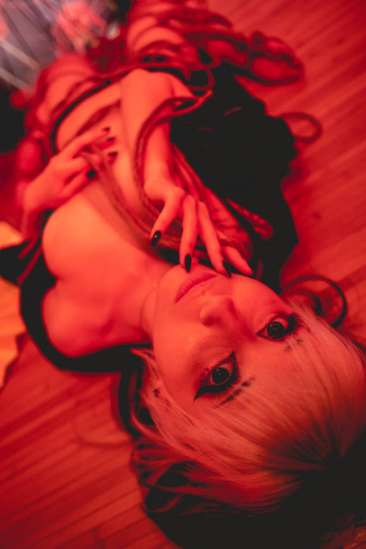 Beautiful, slim, young and sexy girl with blonde locks, small and sexy black kimono and panties, and shibari ties and knots on the body, in a traditional japanese room with red light - Foto, afbeelding
