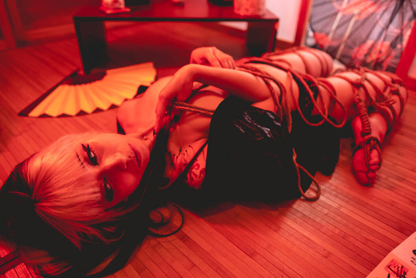 Beautiful, slim, young and sexy girl with blonde locks, small and sexy black kimono and panties, and shibari ties and knots on the body, in a traditional japanese room with red light - Foto, imagen