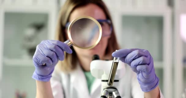 Chemist examines test tube with dark liquid or crude oil through magnifying glass. Chemical laboratory equipment toxic and poisonous substances - Filmagem, Vídeo