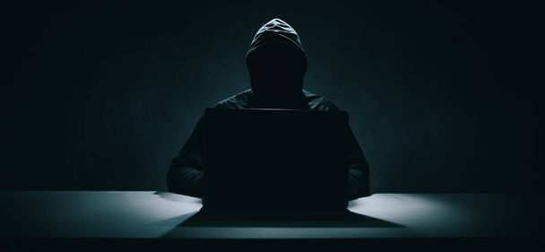 Хакер в черной толстовке работает над ноутбуком в темной комнате. Кибербезопасность. banner - Фото, изображение