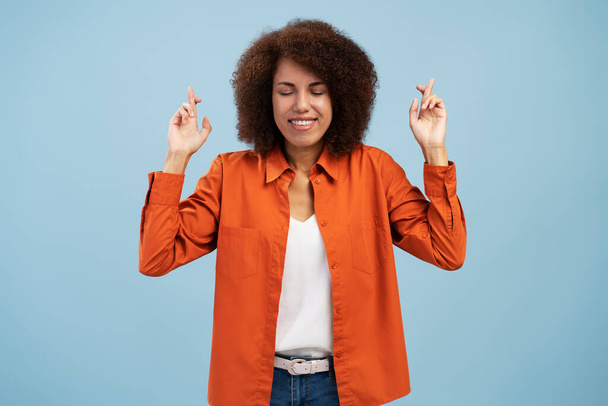 Όμορφη κομψή και εντυπωσιακή Αφροαμερικανή γυναίκα που φοράει πορτοκαλί casual πουκάμισο και μπλε τζιν, κλείνει τα μάτια της και σταυρώνει το δάχτυλο, εκφράζοντας θετικότητα και αισιοδοξία, απομονωμένη σε μπλε φόντο - Φωτογραφία, εικόνα