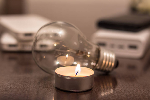 A burning candle against the background of a light bulb and power banks. Blackout due to war in Ukraine - Foto, immagini