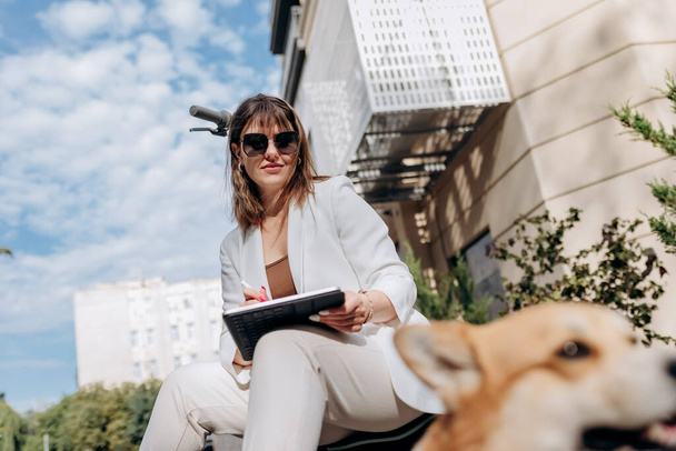 Young businesswoman in white suit sitting on electric scooter and working on digital tablet during walking with Welsh Corgi Pembroke dog in city - Photo, image