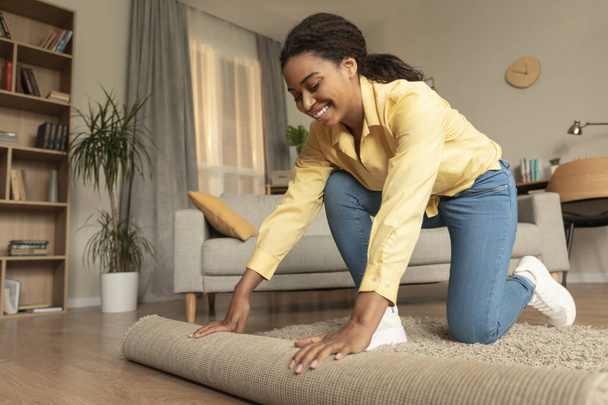 Young african american woman doing the housekeeping, bending down and rolling carpet in a living room, preparing to cleaning the floor, copy space - Foto, immagini