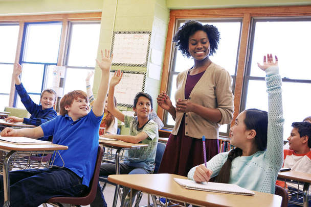 Koulu, ohjaaja ja opiskelijat nostavat kätensä kysymään tai vastaamaan akateemiseen kysymykseen oppimiseen. Monimuotoisuus, koulutus ja peruskoulun lapset puhuvat naisopettajalleen luokkahuoneessa - Valokuva, kuva