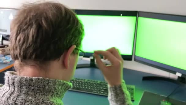 男は2台のコンピュータでオフィスで働いています緑の画面クロマキー彼は後ろに座って、画面を見てメガネを脱いで戻るグレーの部屋赤い髪のプログラマーデザイナー3Dアニメータオフィスの仕事 - 映像、動画